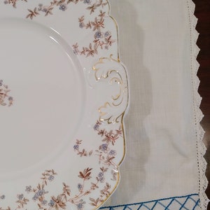 May include: A white porcelain serving platter with a square shape and scalloped edges. The platter has a floral design with brown and blue flowers and gold accents. The platter has two handles on the side.