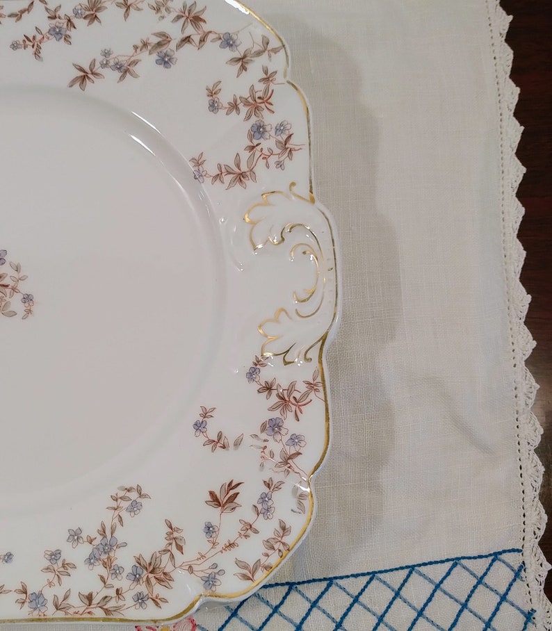 May include: A white porcelain serving platter with a square shape and scalloped edges. The platter has a floral design with brown and blue flowers and gold accents. The platter has two handles on the side.