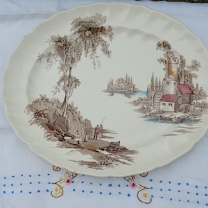 Puede incluir: Un plato de servir ovalado vintage con una escena de paisaje marrón y blanco. La escena presenta un río, un molino de agua, una casa, árboles y dos figuras pescando. El plato tiene un borde festoneado.