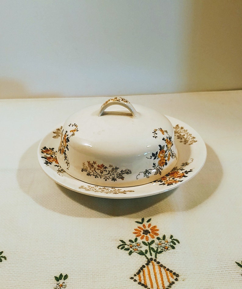 Puede incluir: Un mantequillero vintage blanco con un dise&ntilde;o floral en tonos marrones, amarillos y negros. El mantequillero tiene una tapa con asa y se asienta sobre un plato a juego.