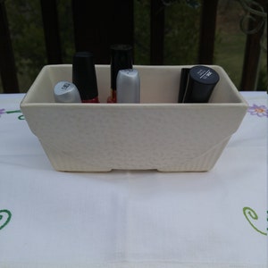 Puede incluir: Un macetero de cerámica blanca con una superficie texturizada contiene una colección de botellas de esmalte de uñas. Las botellas son de varios colores, incluyendo negro, rojo y plateado.