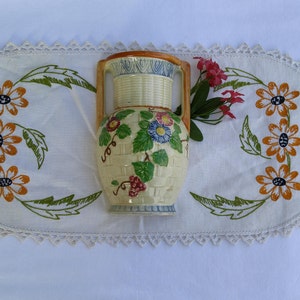 May include: A vintage ceramic vase with a woven pattern and floral designs in shades of yellow, green, blue, and red. The vase has two handles and a rounded base.