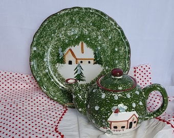 May include: A green and white ceramic teapot with a snowy winter scene and a red lid. The teapot is sitting on a white surface with a red and white polka dot tablecloth.