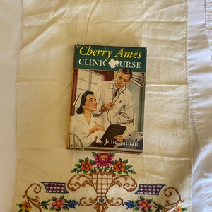 May include: A vintage paperback book titled "Cherry Ames Clinic Nurse" by Julie Tatham. The cover features a nurse and a doctor in a hospital setting.