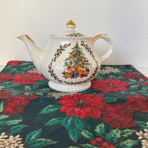 May include: White ceramic teapot with a Christmas tree design. The teapot has gold trim and a lid with a gold finial. The Christmas tree is decorated with colourful ornaments and surrounded by a garland of red berries and green leaves.