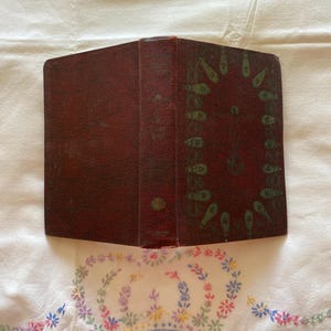 Pode incluir: Um livro antigo encadernado em couro vermelho escuro com um design verde e dourado na capa. O design apresenta um padrão repetitivo de peixes estilizados.
