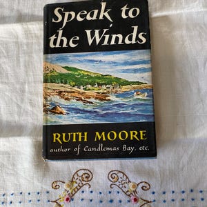 Könnte beinhalten: Ein Vintage-Buch mit dem Titel "Speak to the Winds" von Ruth Moore. Das Cover zeigt eine Küstenlandschaft mit blauem Himmel und Meer. Das Buch liegt auf einem weißen Tuch mit gestickten Blumenmustern.