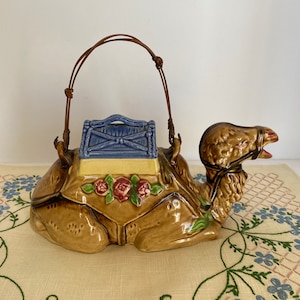 May include: A ceramic camel teapot with a blue and yellow decorated saddle and a brown woven handle. The camel is decorated with brown, yellow, and green floral details.