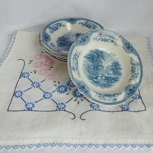 May include: A set of six white and blue ceramic bowls with a landscape design. The bowls are stacked on a white embroidered cloth with pink and blue floral patterns. The bowls have a diameter of approximately 6 inches.