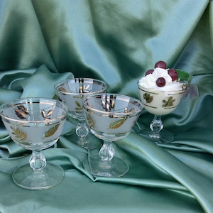 May include: Four clear glass dessert dishes with gold leaf accents and a frosted finish. The dishes are on a green satin fabric background. One dish is filled with whipped cream and red berries.