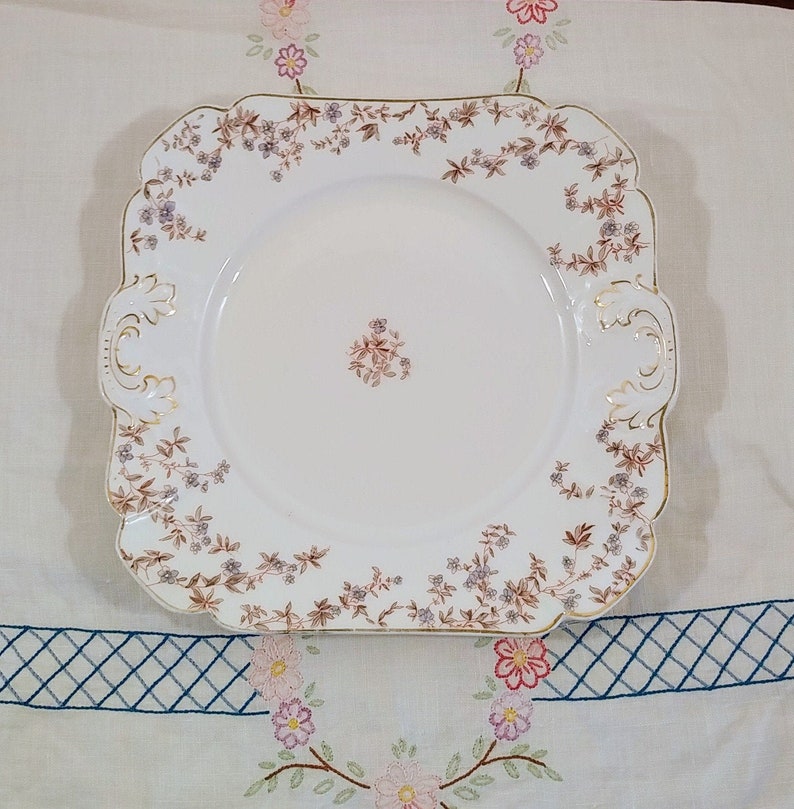 May include: A white square ceramic serving plate with a gold rim and a floral pattern in shades of brown and purple.