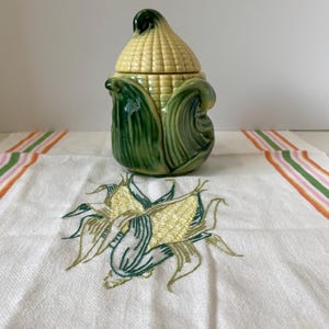 Peut inclure: Un pot en céramique avec couvercle en forme d'épi de maïs avec des feuilles vertes, posé sur un tissu blanc brodé représentant deux épis de maïs avec des feuilles vertes et des bordures rayées orange, rose et vert sur les côtés.