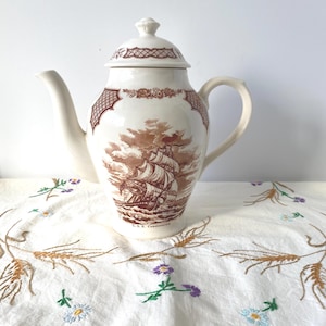 Puede incluir: Tetera de cerámica color crema con tapa, con una ilustración marrón de un velero y patrones decorativos. La tetera tiene un pico y un asa curvados. El texto "U.S.S. Constitution" está debajo.