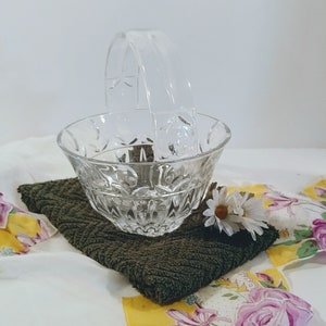 Könnte beinhalten: Ein klarer Glaskorb mit Henkel, mit einem geschliffenen Design. Der Korb steht auf einem dunkelgrünen, strukturierten Tuch, mit einem weißen und floral gemusterten Stoff im Hintergrund. Eine kleine weiße Gänseblümchen ist in der Nähe des Korbes platziert.