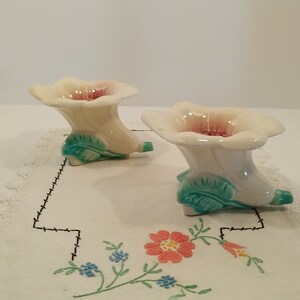 Peut inclure: Deux bougeoirs en céramique blanche en forme de fleur avec des feuilles vertes et des centres roses. Les bougeoirs sont posés sur une nappe blanche brodée.