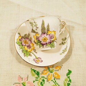 Puede incluir: Un juego de taza y platillo de té vintage con un diseño floral. La taza y el platillo son blancos con un borde dorado. El diseño floral presenta flores amarillas, moradas y verdes.