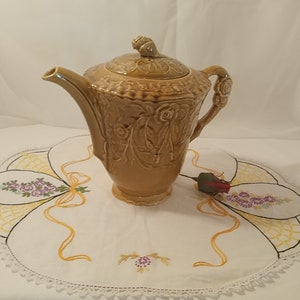 以下が含まれることがあります： 花柄の蓋が付いた茶色の陶器製のコーヒーポット。ポットは側面にハンドル、前面に注ぎ口が付いています。ポットはレースの縁取りが施された白いテーブルクロスの上に置かれています。