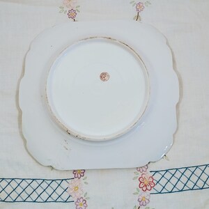 May include: White ceramic plate with a scalloped edge. The plate has a raised center circle and a red stamp on the bottom.