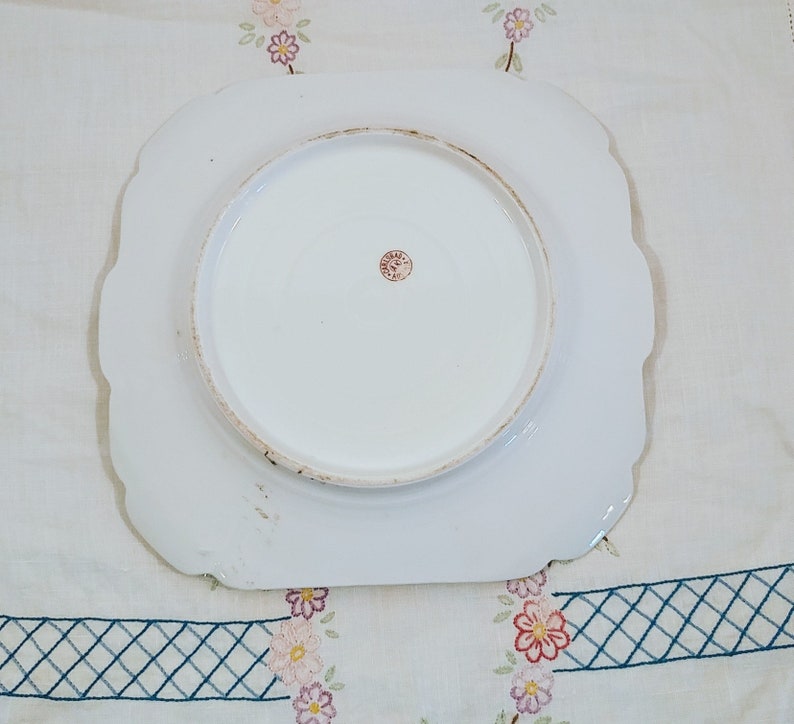 May include: White ceramic plate with a scalloped edge. The plate has a raised center circle and a red stamp on the bottom.