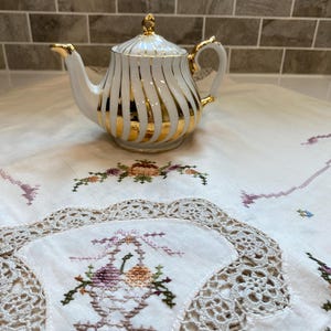 May include: A white and gold porcelain teapot with vertical gold stripes and gold accents. The teapot sits on a white embroidered tablecloth with floral designs. The teapot has a lid and a curved handle.