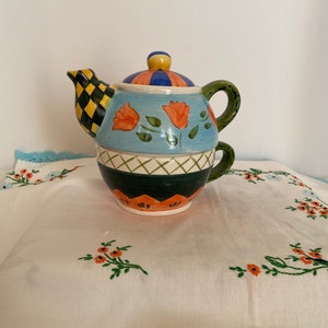 May include: A colorful ceramic teapot and teacup set with a blue, green, orange, and yellow floral design. The teapot has a checkered spout and a striped lid.