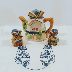May include: A ceramic teapot and two matching salt and pepper shakers shaped like windmills. The teapot is decorated with blue, green, and brown paint and has a white base. The salt and pepper shakers are smaller and have a similar design.