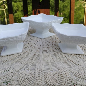 May include: Three white milk glass pedestal bowls with a floral design. The bowls are on a white crocheted doily.