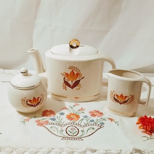 Könnte beinhalten: Ein weißes Keramik-Tee-Set mit floralem Design. Das Set enthält eine Teekanne, eine Milchkännchen und eine Zuckerdose. Die Teekanne hat einen goldenen Rand und ein florales Design mit orangefarbenen und braunen Akzenten. Die Milchkännchen und die Zuckerdose haben ein ähnliches florales Design.