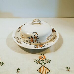Puede incluir: Un mantequillero vintage de cer&aacute;mica blanca con un dise&ntilde;o floral. El mantequillero tiene una tapa con asa y un plato a juego.