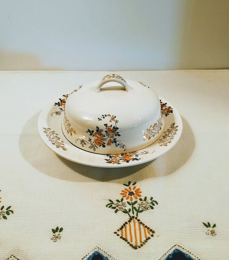 Puede incluir: Un mantequillero vintage de cer&aacute;mica blanca con un dise&ntilde;o floral. El mantequillero tiene una tapa con asa y un plato a juego.