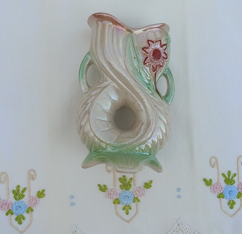 May include: A vintage ceramic vase with a pearlescent glaze and a green and white floral design. The vase has a unique shape with a hole in the center and a red flower with a yellow center.