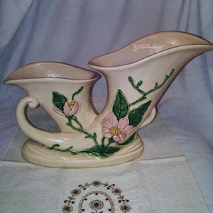 May include: A double-handled ceramic vase with a cream-colored glaze and a floral design. The vase features pink flowers with yellow centers and green leaves.
