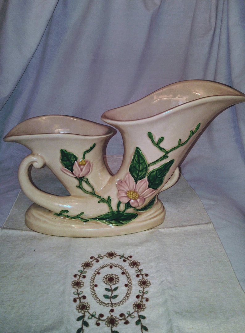 May include: A double-handled ceramic vase with a cream-colored glaze and a floral design. The vase features pink flowers with yellow centers and green leaves.