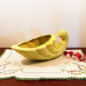 Może przedstawiać: Żółty ceramiczny wazon w kształcie rogu obfitości z zakrzywionym uchwytem. Wazon stoi na białym haftowanym obrusie z zieloną obwódką.