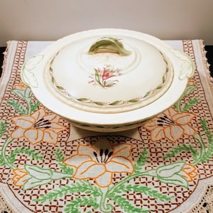 Może przedstawiać: Biała ceramiczna miska do serwowania z pokrywką i wzorem kwiatowym. Miska ma zielony uchwyt i jest ozdobiona zielonymi liśćmi i różowymi kwiatami. Miska stoi na białym obrusie z haftowanymi kwiatami.