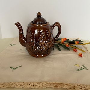 May include: A brown ceramic teapot with a lid and a floral design. The teapot is sitting on a white tablecloth with a crocheted edge.