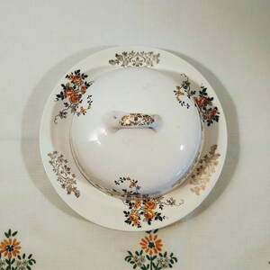 Puede incluir: Un mantequillero de cer&aacute;mica blanca con un dise&ntilde;o floral en tonos naranja, marr&oacute;n y dorado. El mantequillero tiene una tapa con asa.