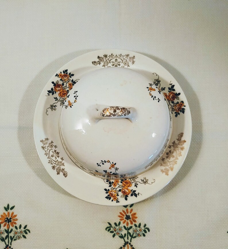 Puede incluir: Un mantequillero de cer&aacute;mica blanca con un dise&ntilde;o floral en tonos naranja, marr&oacute;n y dorado. El mantequillero tiene una tapa con asa.