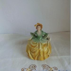 Może przedstawiać: Porcelanowa figurka kobiety w zielono-żółtej sukni. Ma na sobie kapelusz i ma włosy związane do tyłu. Figurka stoi na białym tle.