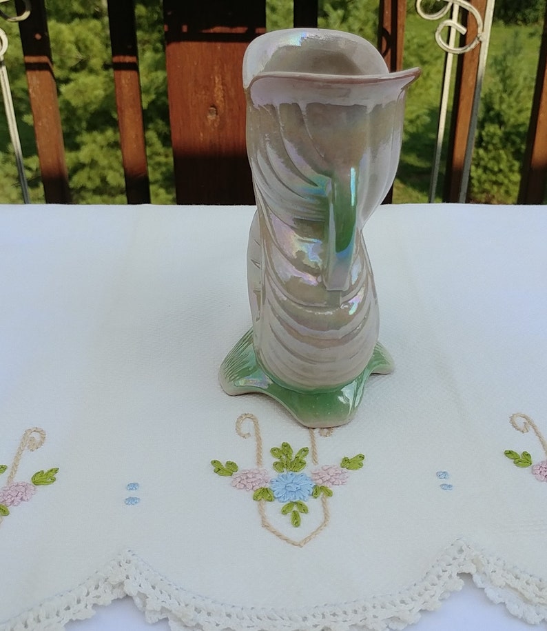 May include: A white ceramic vase with a green base and iridescent glaze. The vase is shaped like a stylized bird with a long neck and a curved body.