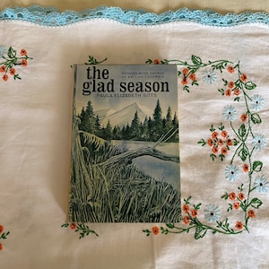 May include: A vintage paperback book titled "the glad season" by Paula Elizabeth Sitts. The book cover features a watercolor illustration of a forest scene with a lake and trees.