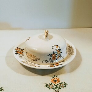 Puede incluir: Un mantequillero vintage blanco con un dise&ntilde;o floral en tonos amarillos, marrones y azules. El mantequillero tiene una tapa con asa y se asienta sobre un plato a juego.