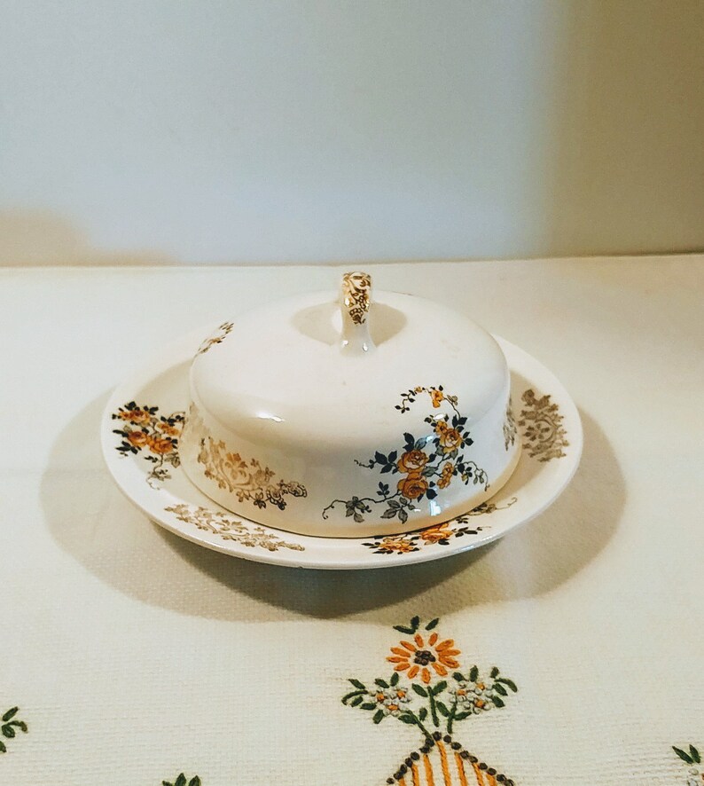 Puede incluir: Un mantequillero vintage blanco con un dise&ntilde;o floral en tonos amarillos, marrones y azules. El mantequillero tiene una tapa con asa y se asienta sobre un plato a juego.