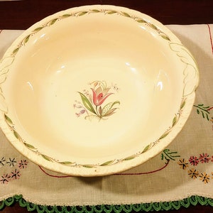 Może przedstawiać: Duża, kremowa ceramiczna miska z zielonym i brązowym wzorem kwiatowym. Miska ma falisty brzeg i dwa uchwyty.