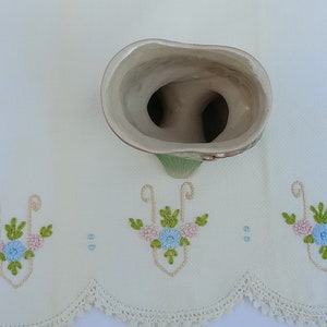 May include: A white ceramic vase with a green and brown floral design. The vase has two openings at the top, and the inside is white.