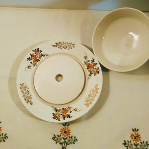 Puede incluir: Un mantequillero de cer&aacute;mica vintage con un dise&ntilde;o floral. El mantequillero tiene una tapa con un peque&ntilde;o agujero en el centro. El mantequillero es blanco con acentos florales naranja y marr&oacute;n.