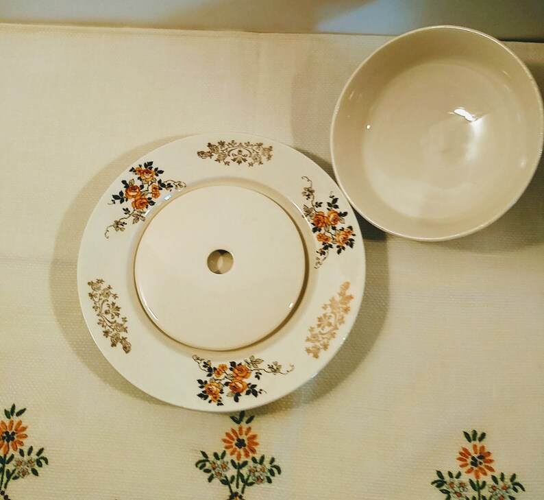 Puede incluir: Un mantequillero de cer&aacute;mica vintage con un dise&ntilde;o floral. El mantequillero tiene una tapa con un peque&ntilde;o agujero en el centro. El mantequillero es blanco con acentos florales naranja y marr&oacute;n.