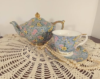 May include: A blue and white floral teapot and teacup set with gold accents. The teapot has a lid and a spout. The teacup has a handle and a saucer. The set is on a white crocheted doily with a pearl necklace.