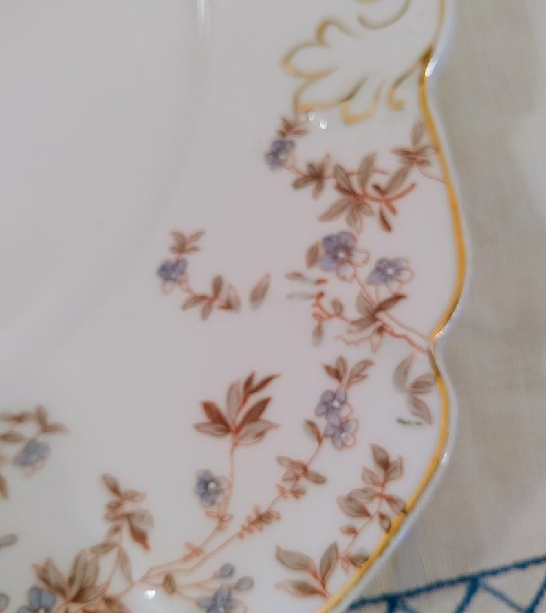 May include: A white porcelain plate with a scalloped edge and a gold trim. The plate is decorated with a delicate floral pattern in shades of brown and blue.