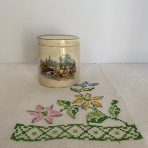 May include: A cream-colored ceramic jar with a lid, featuring a scenic landscape design with trees, water, and figures. The jar is set on a white embroidered cloth with floral cross-stitch patterns.